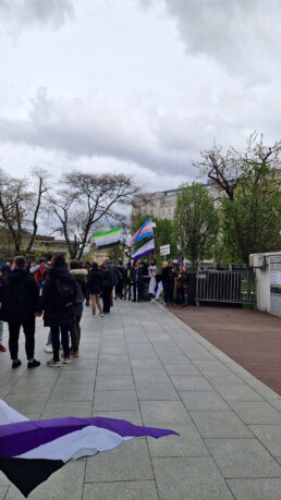 Gyülekező felvonulók a budapesti Erzsébet téren. A csoport egy aromantikus-, egy transz- és egy aszexuális zászlót emel a magasba. Mellettük egy felvonuló "Támogató szülők csoportja" táblát tart.