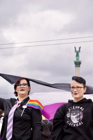 Két sötét ruhás, szivárványos arcfestésű felvonuló egy aszexuális zászlót lenget a budapesti Hősök terén. Egyikőjük pulóverén egy aszexuális-zászló-színű dinoszaurusz van, "Asexual T-Rexual" felirattal.