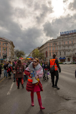 Sulyok Viktória szervező a menet elején megafonnal a kezében a budapesti Oktogonnál, mögötte a felvonulók szivárványos esernyőkkel.