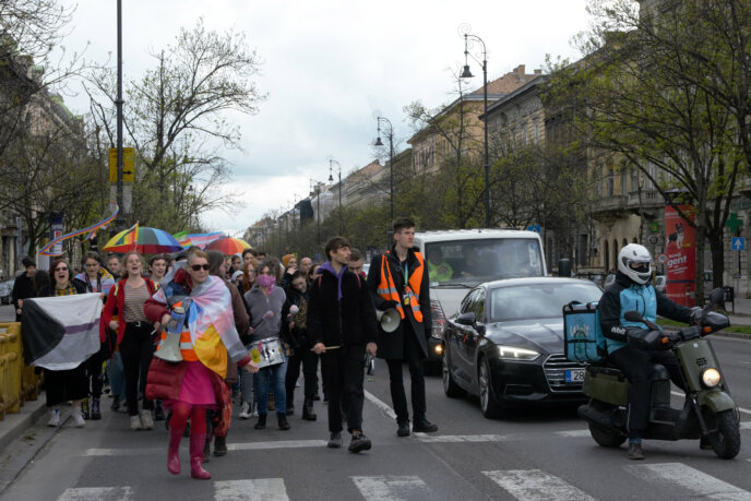A felvonuló menet eleje egy Budapesti úton, mellettük a lámpánál várakozó kocsisor. Többen különböző Pride zászlókat és esernyőket tartanak.