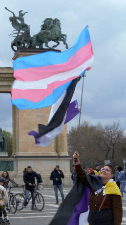 Egy nem-bináris zászlót köpenyként viselő, mosolygó felvonuló egy transz- és egy aszexuális zászlót emel a magasba a budapesti Hősök terén.