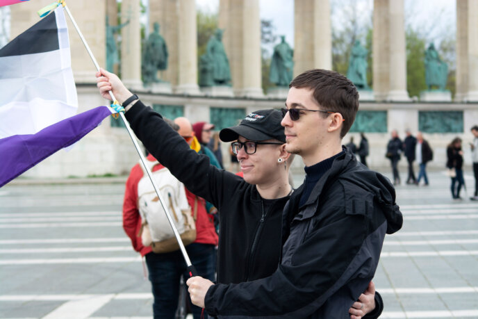 Két sötét ruhába öltözött felvonuló egymást átkarolva egy aszexuális zászlót tart a budapesti Hősök terén.